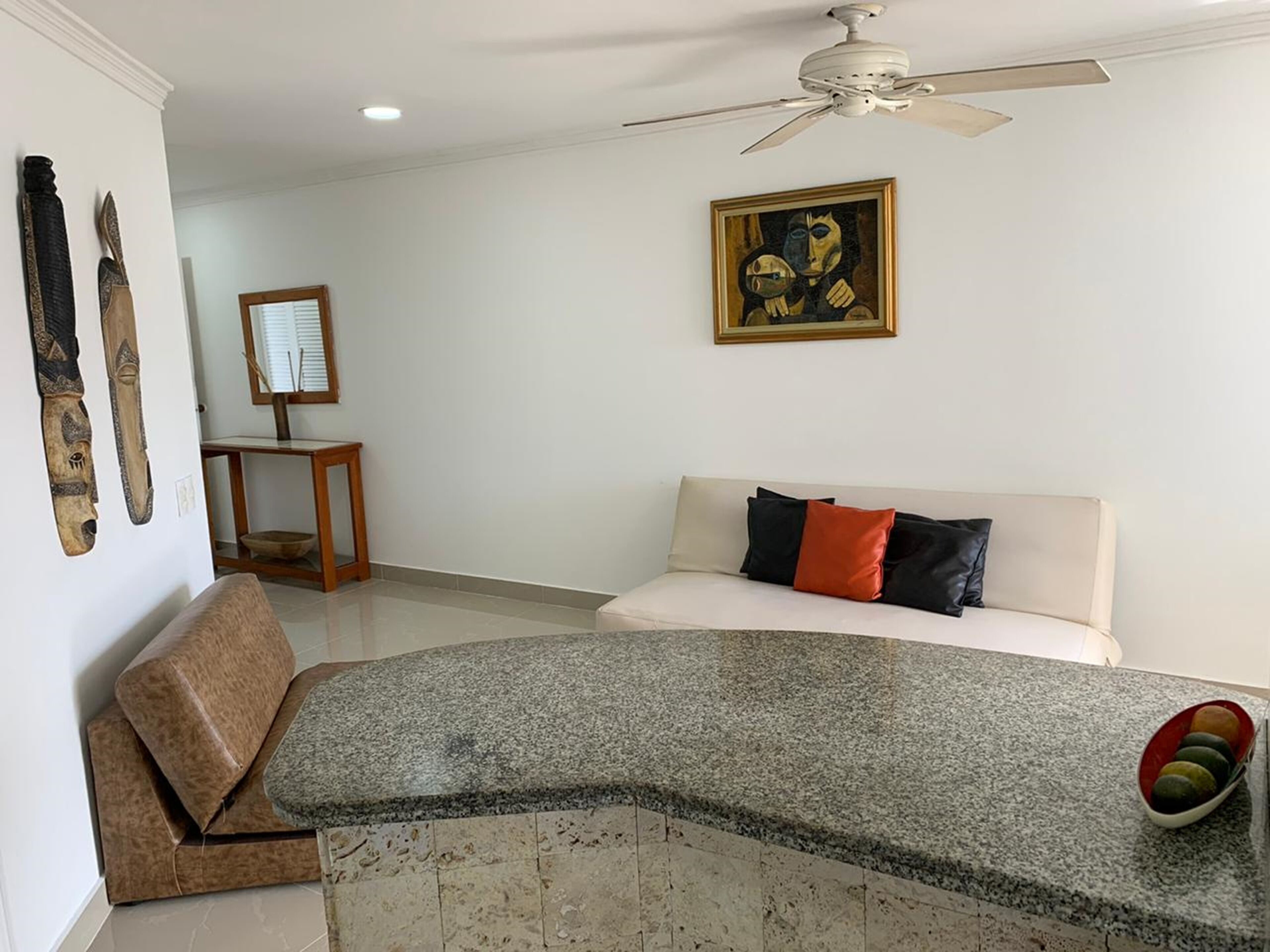Quarto de hotel moderno com bancada de granito curva, cama com várias almofadas, ventoinha no teto e um quadro na parede.