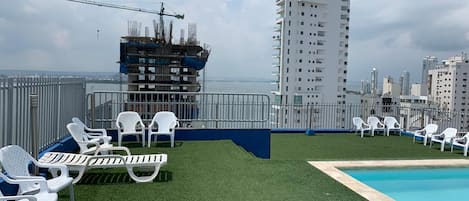 A rooftop pool area with white lounge chairs and a pool, surrounded by a high-rise building under construction.