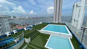 Ein Dachpool mit zwei Bereichen, umgeben von weißen Liegestühlen und Blick auf Meer und Stadtlandschaft.