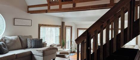A spacious living area with a high wooden ceiling, a staircase, and a large window providing natural light.