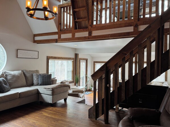 A spacious living area with a high wooden ceiling, a staircase, and a large window providing natural light. - Relax near Gettysburg ski/wineries/fire pit! (Orrtanna)