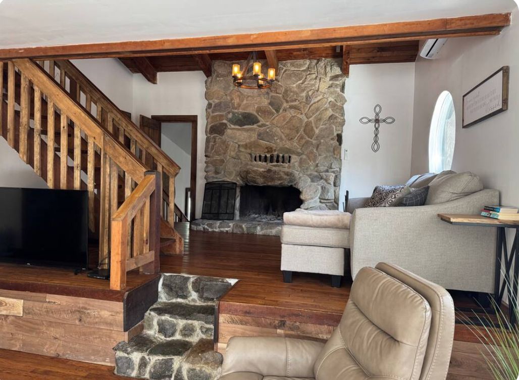 A living room with a stone fireplace, wooden staircase, and a television.