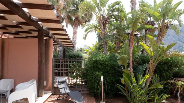 Un patio couvert avec deux chaises blanches, un barbecue en métal et diverses plantes tropicales.