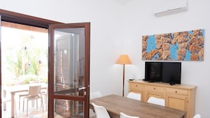 Une pièce comprenant une table à manger en bois, des chaises blanches, un téléviseur posé sur un meuble en bois, un tableau accroché au mur et une porte-fenêtre coulissante donnant sur un espace extérieur aménagé avec des chaises et une table.