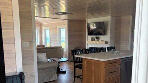 A cozy cabin interior with a wooden floor, a sofa, a dining table with chairs, a kitchen area with a TV, and a large window overlooking a scenic view. - Saltwater Song Beach House (New London)