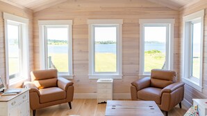 A cabin with wooden walls, three large windows, two leather chairs, a wooden table, and a small white dresser. - Saltwater Song Beach House (New London)