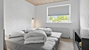 Une chambre avec des murs en briques blanches, un plafond en bois, une fenêtre avec des stores, un lit avec des oreillers et une couverture, et un radiateur.