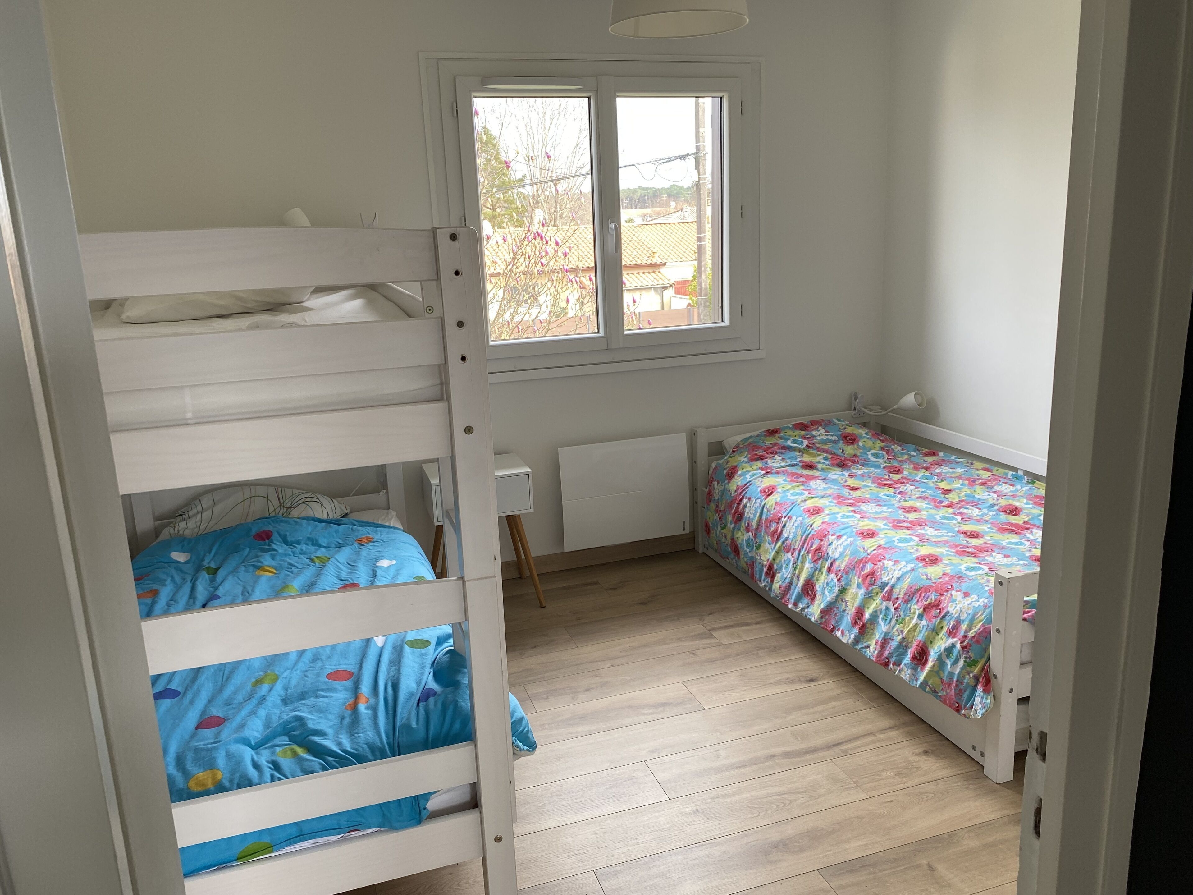 Une chambre avec un lit superposé, un lit simple recouvert d’une courtepointe à motifs floraux, une fenêtre et un radiateur.