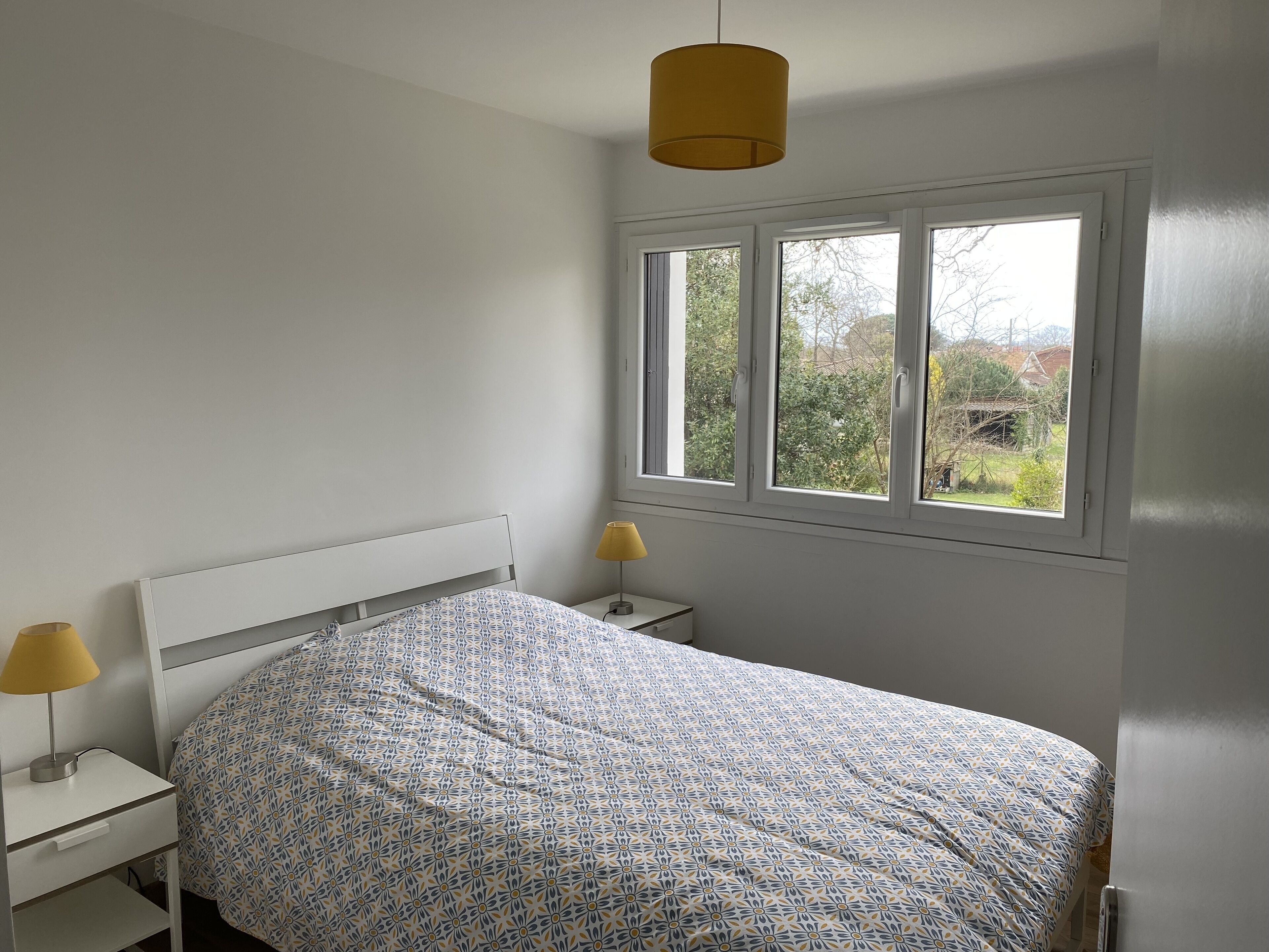 A bedroom with a bed, two bedside tables, a window, and a hanging light fixture.