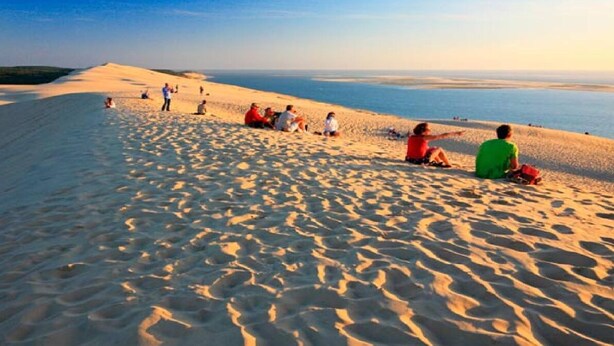 A sandy beach with people sitting and standing, overlooking a calm sea.