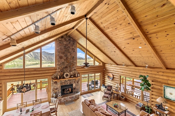 A spacious log cabin interior with a stone fireplace, large windows, and a deck overlooking a scenic landscape.