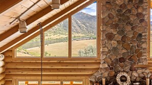 Ein Holzblockhaus-Interieur mit Kamin aus Stein, großen Fenstern mit Blick auf eine hügelige Landschaft und Holzbalken.