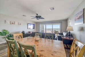 A dining area with a wooden table and chairs, a ceiling fan, and a view of the outdoors through a large window. - Four Shores #3 Gulf Condo Direct Gulf w Pool & Spa (Indian Shores)