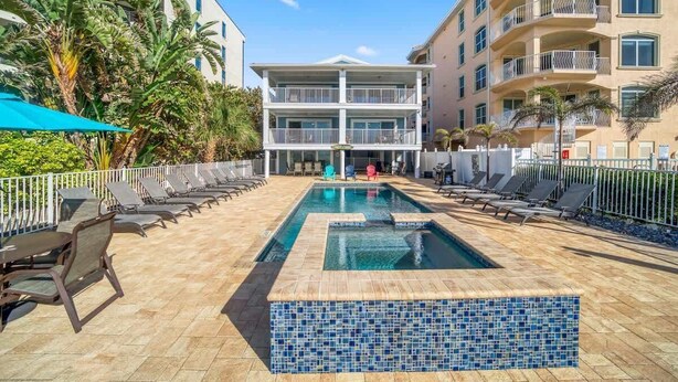 A hotel pool area with lounge chairs, a tiled pool, and a two-story building with balconies.