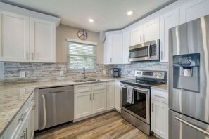 A modern kitchen with white cabinets, stainless steel appliances, a stone backsplash, and a window with blinds. - Four Shores #3 Gulf Condo Direct Gulf w Pool & Spa (Indian Shores)