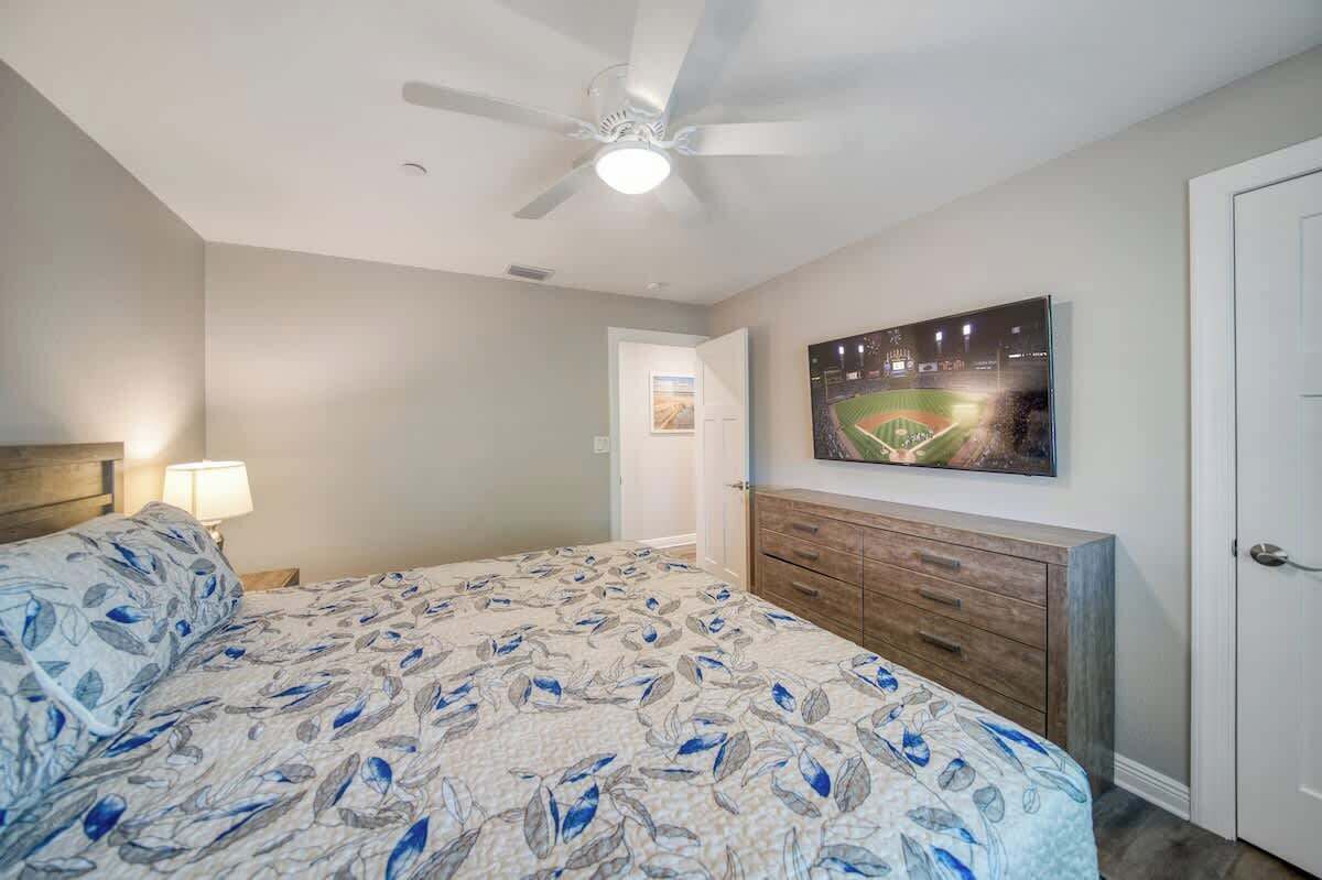 Un dormitorio con cama, ventilador de techo, mesita de noche, cómoda y un cuadro en la pared.