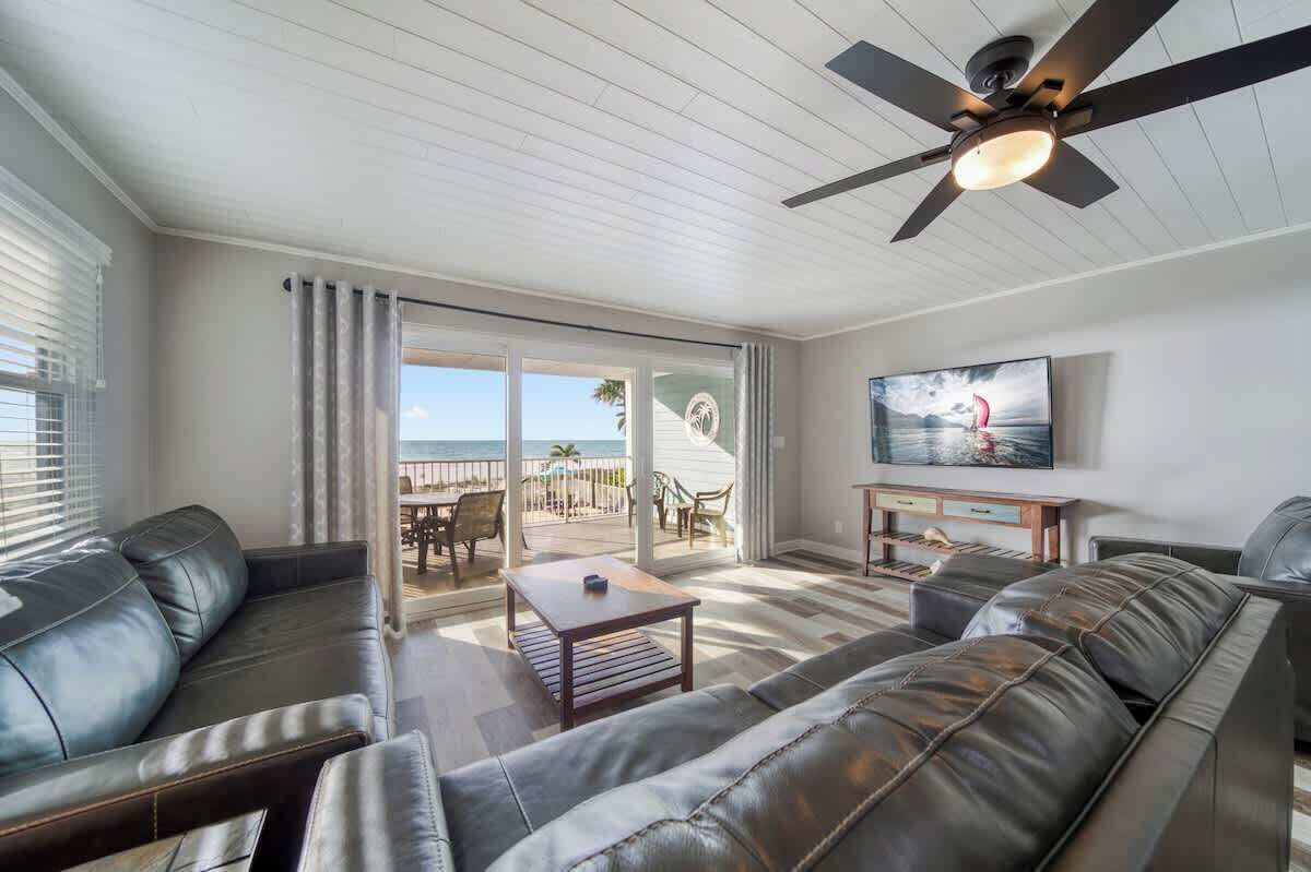 Ein Wohnzimmer mit Deckenventilator, Fernseher und Blick auf den Strand durch große Schiebetüren.