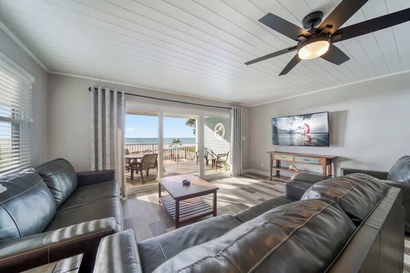 A living room with a ceiling fan, a TV, and a view of the beach through large sliding doors.