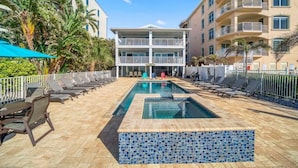 A hotel pool area with lounge chairs, a tiled pool, and a building with balconies. - Four Shores #2 Sunset Condo Direct Gulf w Pool&Spa (Indian Shores)