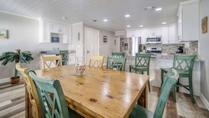 A dining area with a wooden table and chairs, a kitchen with white cabinets, and a potted plant. - Four Shores #2 Sunset Condo Direct Gulf w Pool&Spa (Indian Shores)