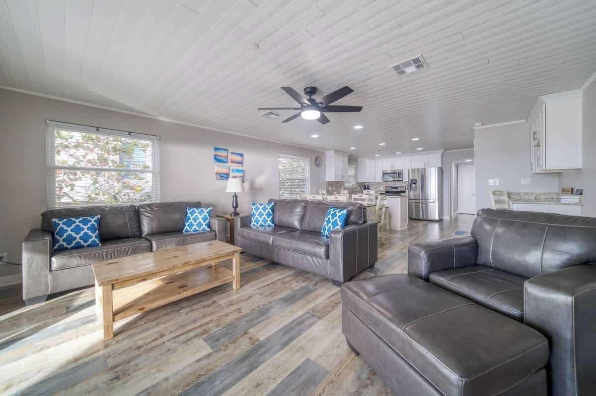 A spacious living area with a wooden coffee table, two leather sofas with patterned cushions, a ceiling fan, and a kitchen in the background.