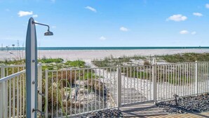 Une vue sur la plage avec une clôture en métal, une terrasse en bois et une structure métallique, qui pourrait être une douche, donnant sur l’océan.