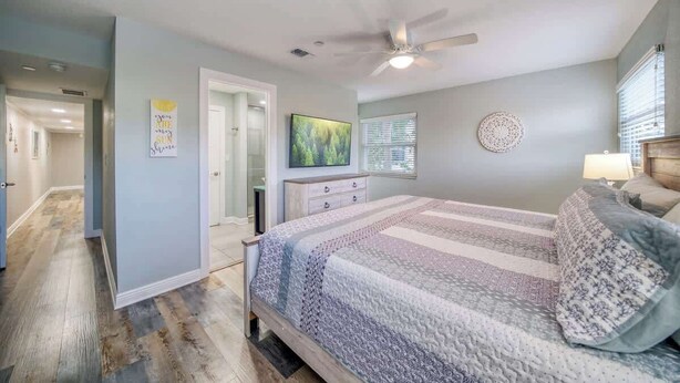 Une chambre à coucher comprenant un lit, un ventilateur de plafond, une table de chevet, une commode et une fenêtre avec des stores.