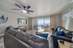 A spacious living area with a ceiling fan, a view of the ocean through large windows, and a balcony with outdoor furniture.