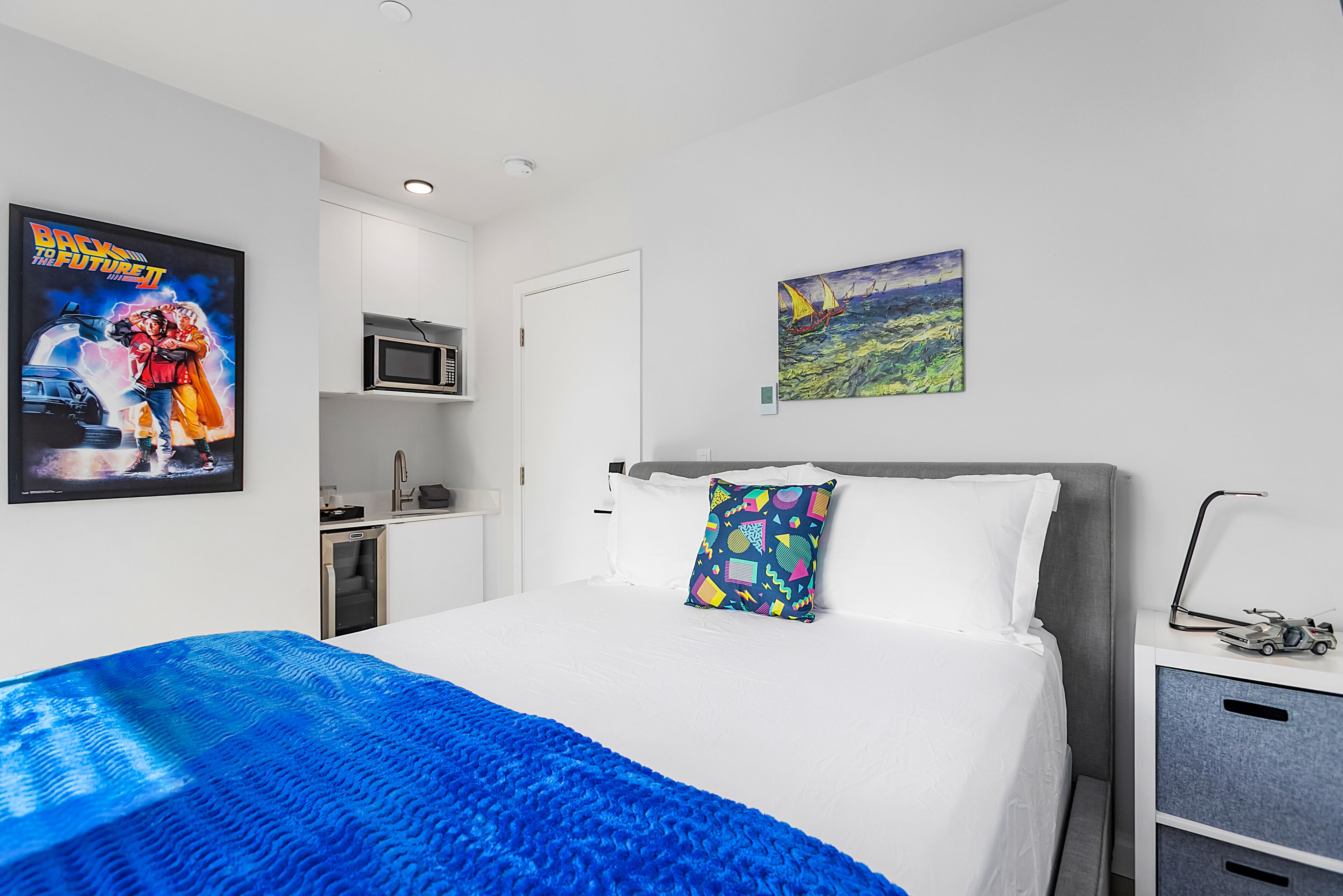 A hotel room with a bed, a microwave, a painting, and a poster on the wall.
