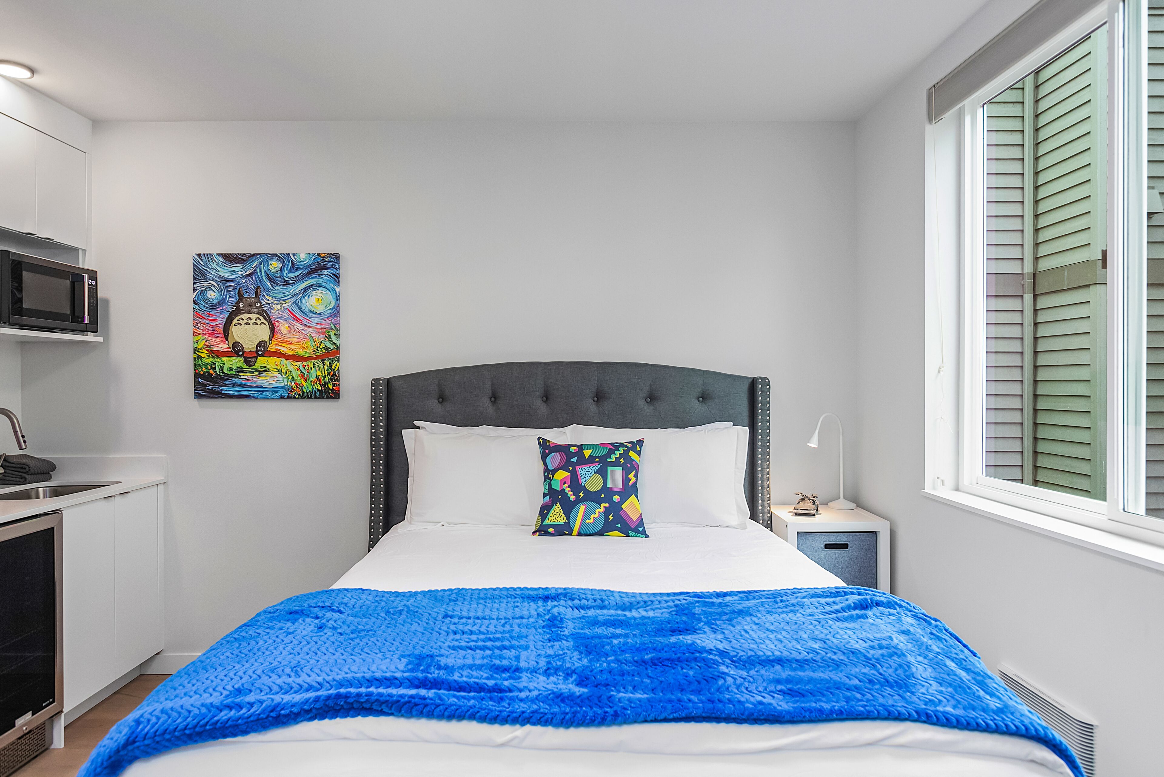 Une chambre à coucher avec un lit, un tableau coloré, une table de chevet et une fenêtre avec des volets.