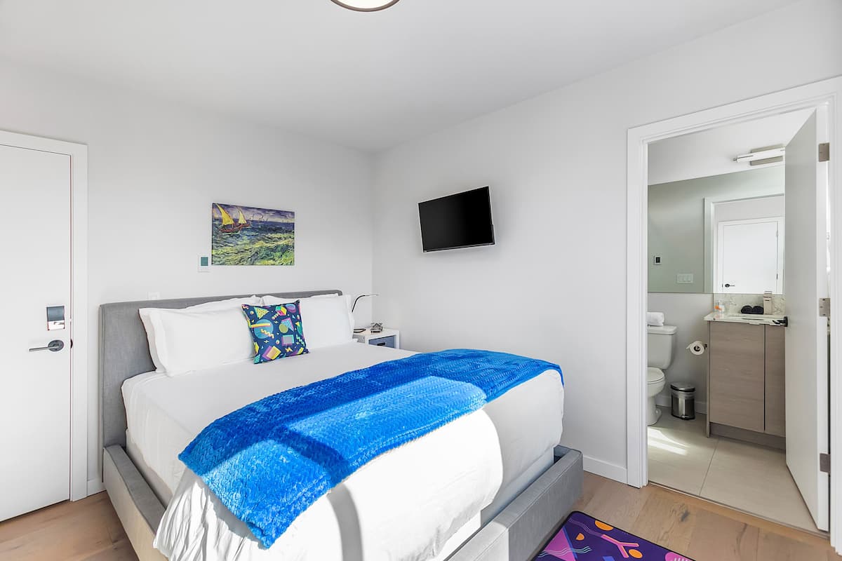 A modern bedroom with a large bed, a flat-screen TV, a painting, and a bathroom visible through an open door.