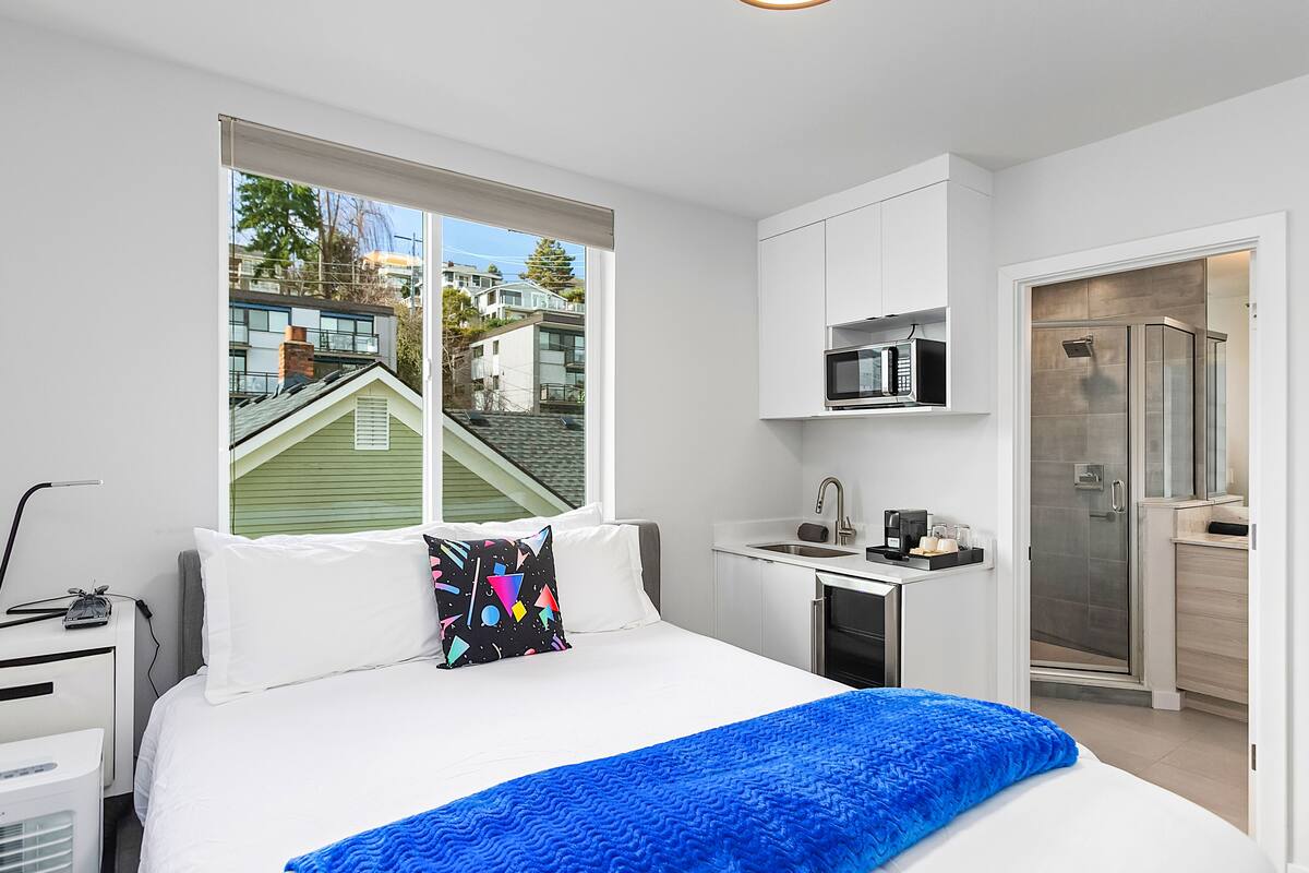 A modern bedroom with a bed, a desk, a microwave, and a shower.