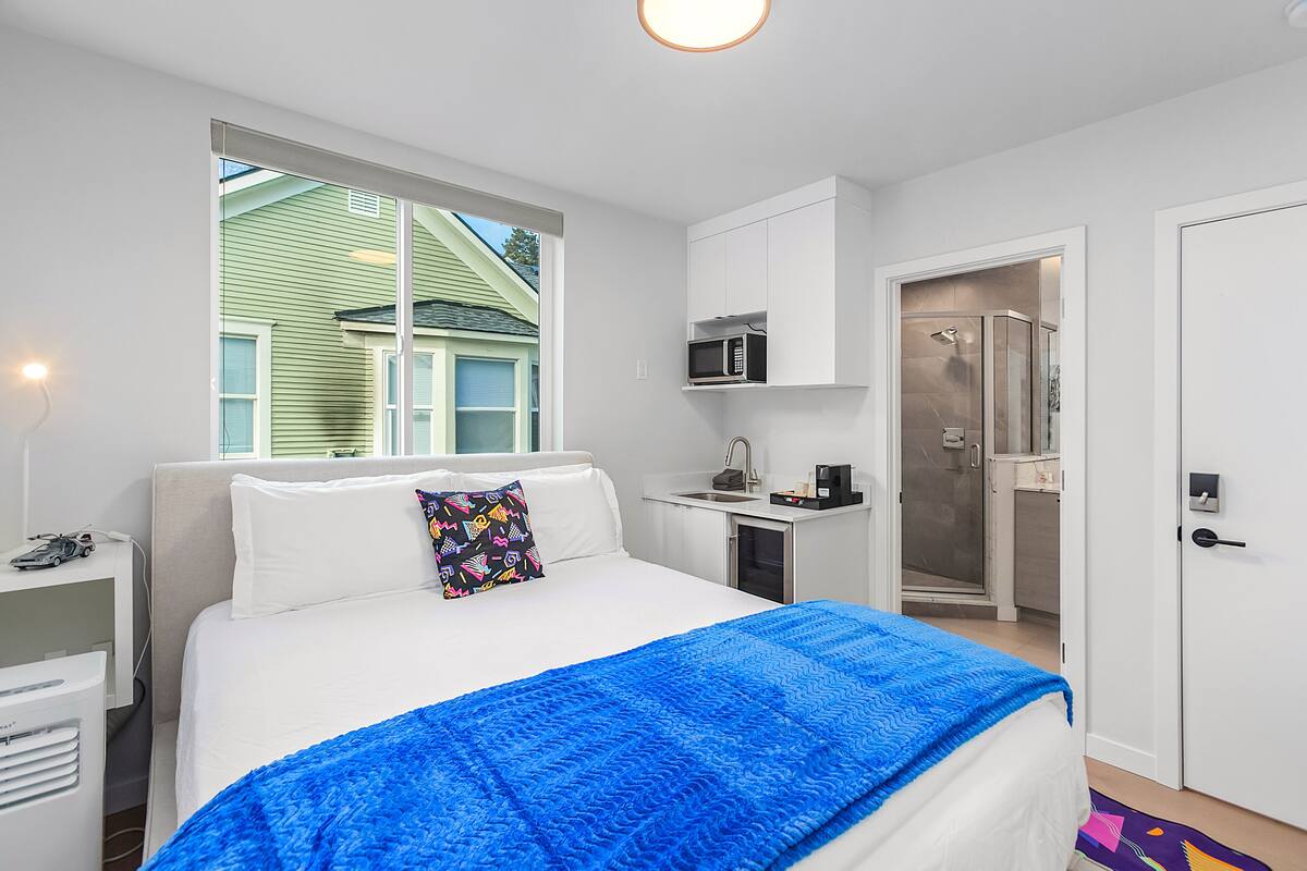 A bedroom with a bed, a window, a microwave, a shower, and a desk.