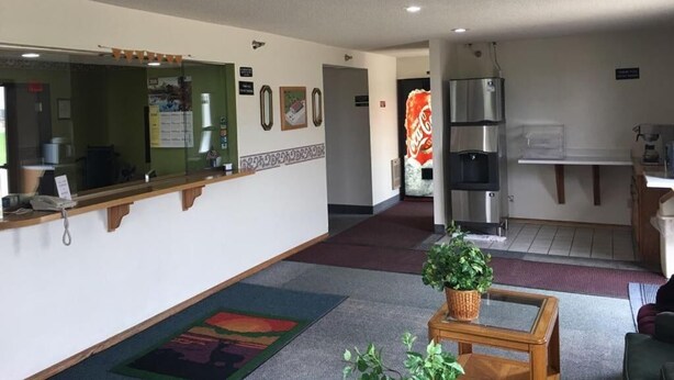 A hotel lobby with a reception desk, a coffee machine, a refrigerator, and a small seating area with a plant. - Prairie Rose inn   (Edgeley)