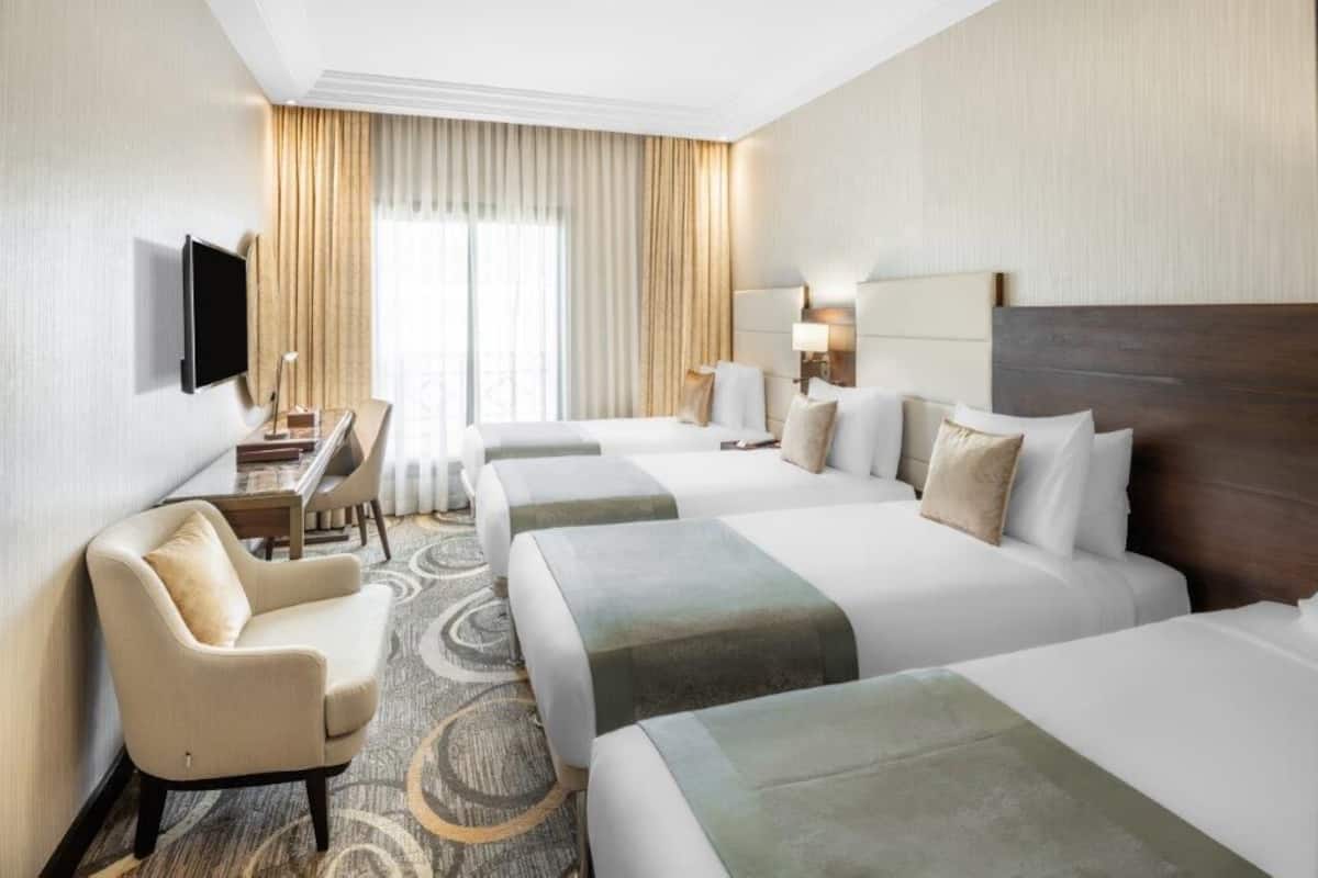 A hotel room with three beds, a desk, a chair, a TV, and a large window with curtains.