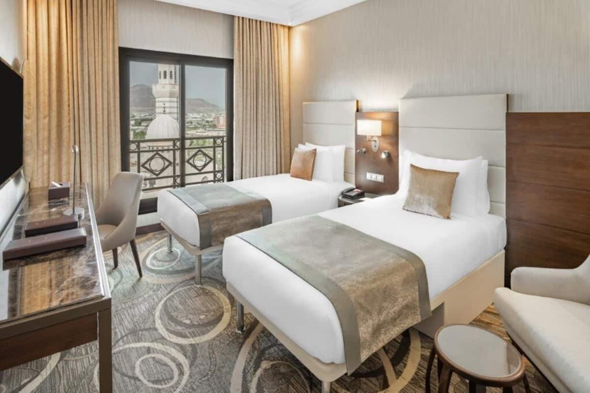 A hotel room with two beds, a desk, a chair, and a view of a cityscape.