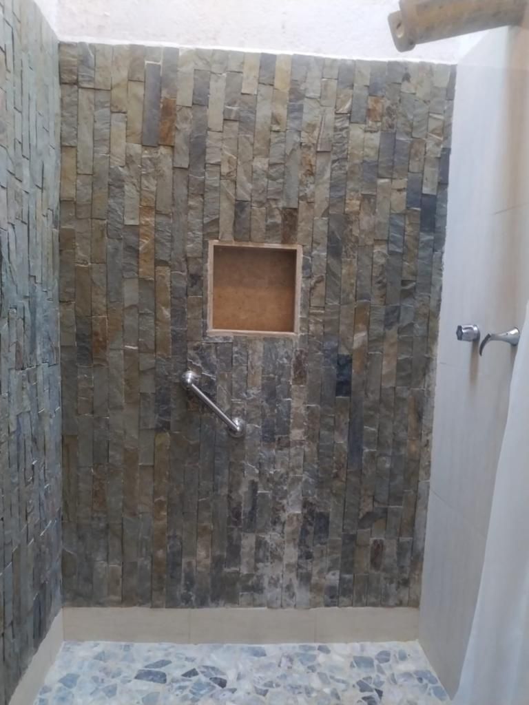 Zona de ducha con pared de piedra, un pequeño estante de madera y un cabezal de ducha.