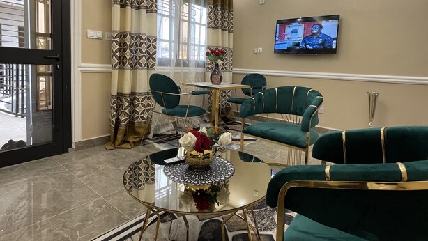 A modern living room with a round glass coffee table, green upholstered chairs, a television on the wall, and a patterned curtain. - JPN SQUARE Douala (Douala)