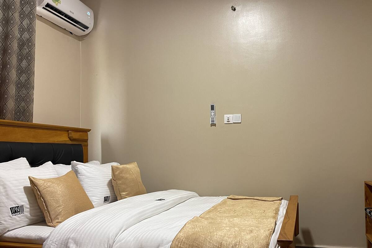 A hotel room with a bed, a bedside table, an ironing board, a wall-mounted air conditioner, and a small wooden cabinet.