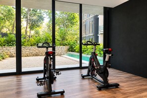 A room with two stationary bikes, large windows with a view of greenery and a building, and a wooden floor.