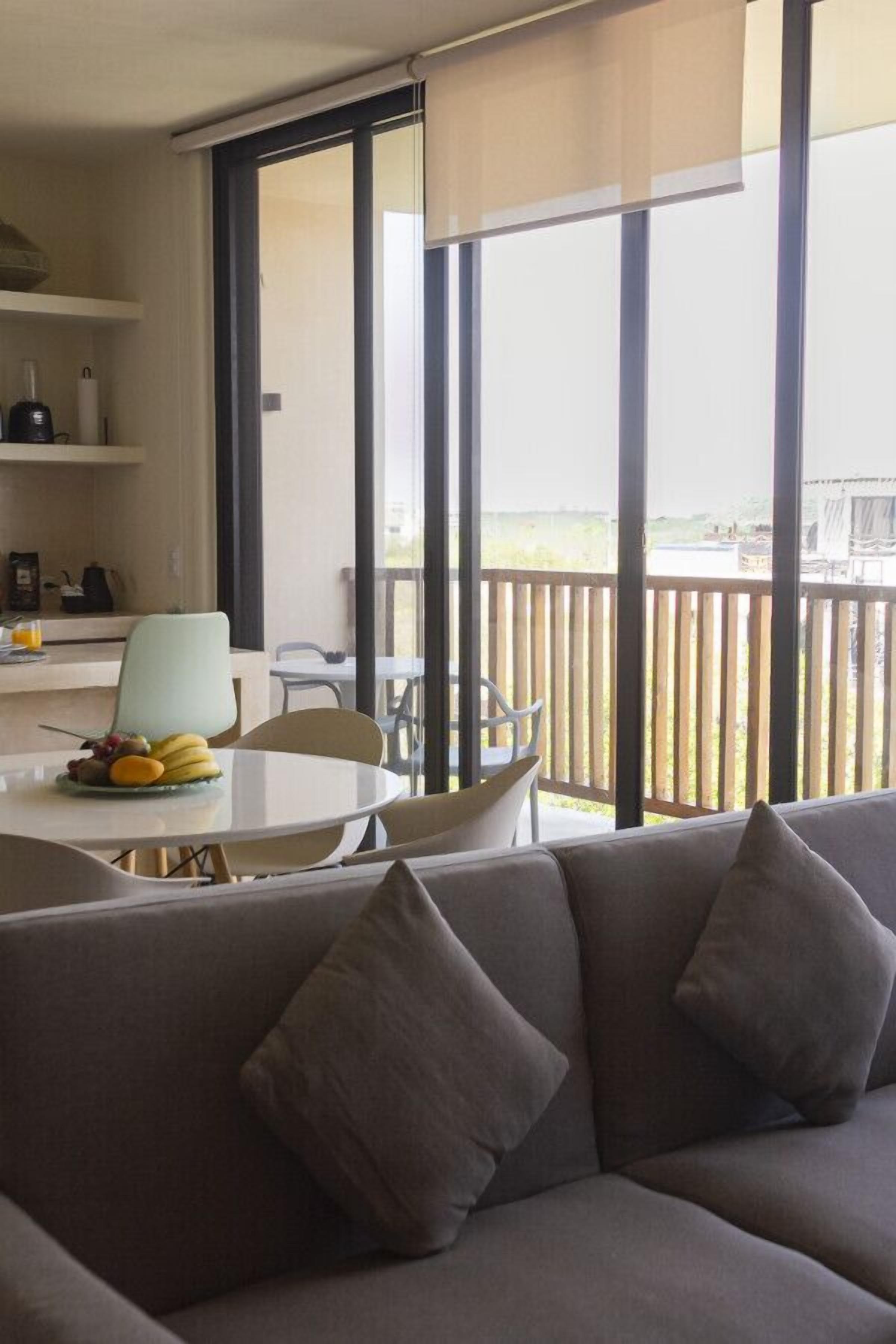 A modern living room with a grey sofa, a dining table with fruit, and a balcony with a wooden railing.