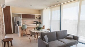 Una cocina moderna con refrigerador, estufa y lavaplatos. Hay una mesa de comedor con sillas y un sofá en una habitación luminosa con ventanas amplias.