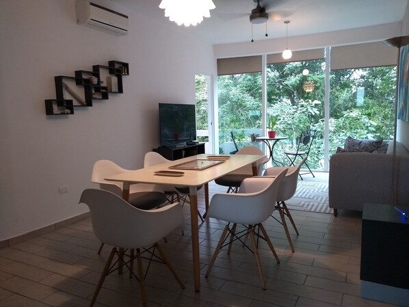 A modern dining area with a wooden table and white chairs, a flat-screen TV, and a large window offering a view of greenery.
