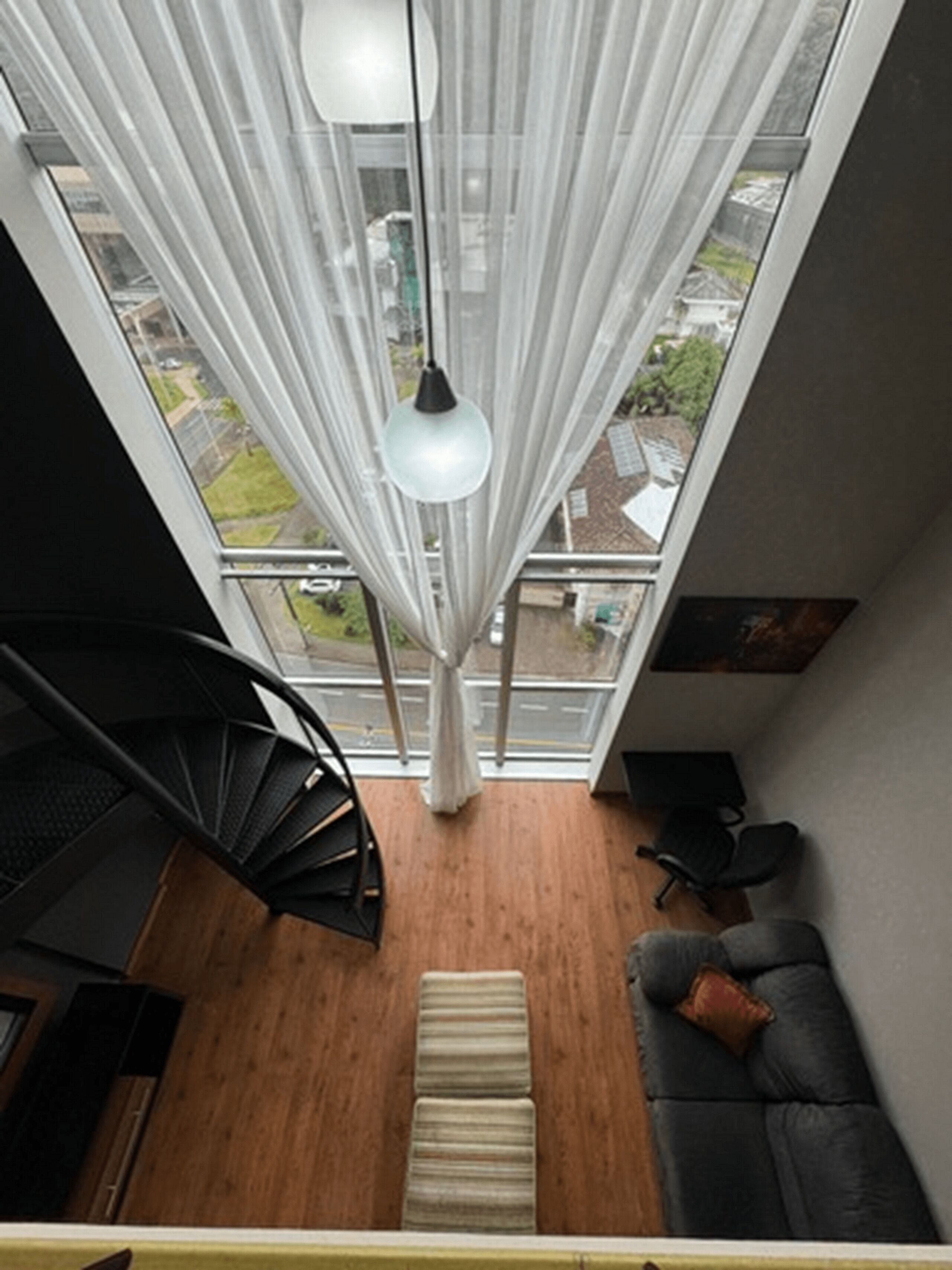 Un interior moderno con una escalera espiral, un sofá gris con un cojín, una lámpara colgante y vistas amplias a través de grandes ventanales.