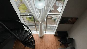 Un interior moderno con una escalera espiral, un sofá gris con un cojín, una lámpara colgante y vistas amplias a través de grandes ventanales.
