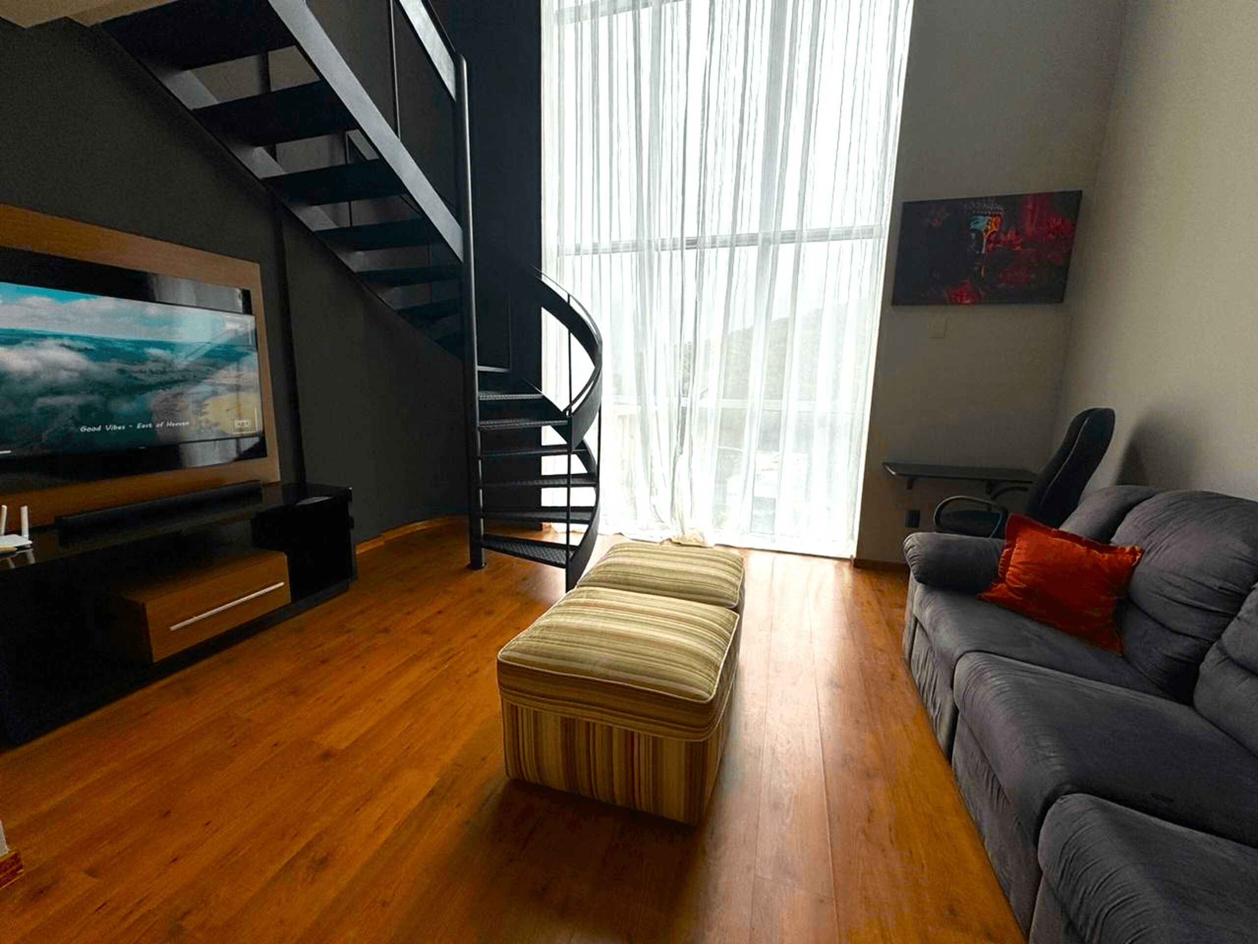 A modern living room with a flat-screen TV, a grey sofa, a striped ottoman, and a staircase.