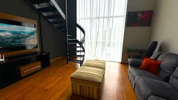 A modern living room with a flat-screen TV, a grey sofa, a striped ottoman, and a staircase.