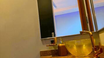 Un baño con un espejo grande, un lavabo con lavamanos dorado y un dispensador de papel higiénico en la pared.