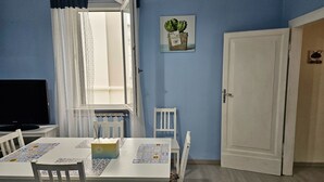 Et værelse med et spisebord, stole, et fjernsyn, et vindue med gardiner, en dør og et billede hængt på væggen.
