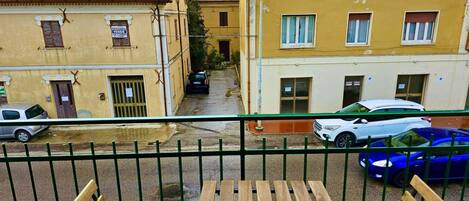 A balcony with a view of a narrow street lined with yellow buildings, a parked car, and a blue car.